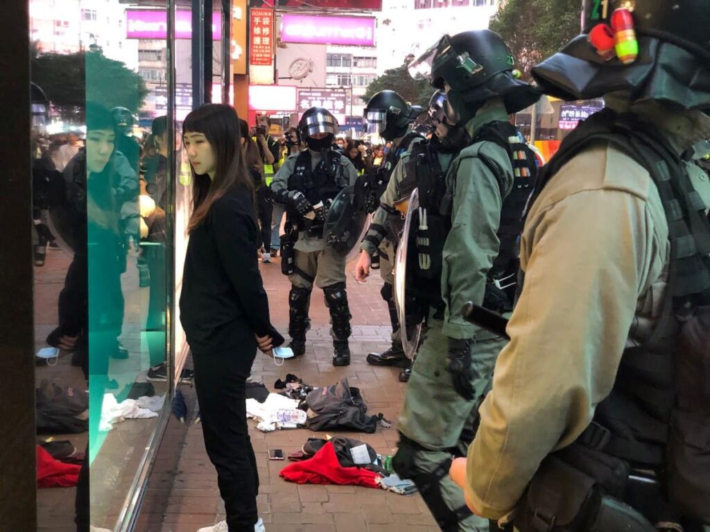 A woman dressed in black looks at her reflection in a shop window, surrounded by officers in riot gear