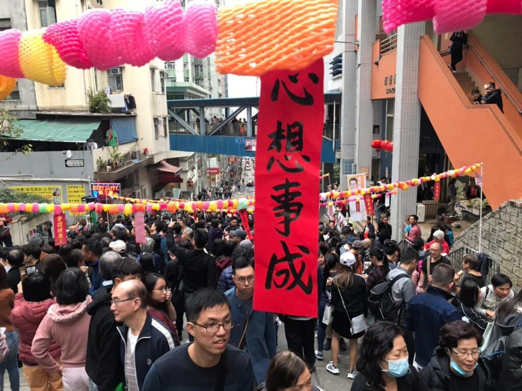 Milling crowds in a Hong Kong street with holiday decorations of a vertical red banner with Chinese writing and colorful paper chains stretched across the street