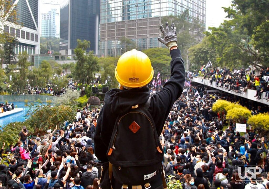 A protestor in a yellow hard hat and a backpack, facing away from the camera and holding up a gloved hand towards a large crowd of protestors below