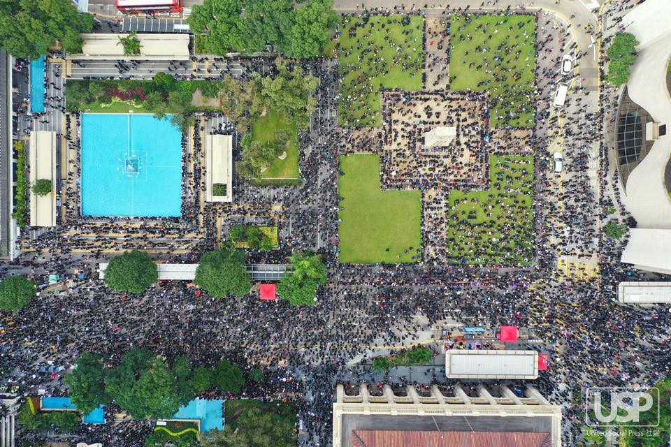 An aerial view of a square, including fountains, geometrical lawns, and trees, full of thousands of protestors