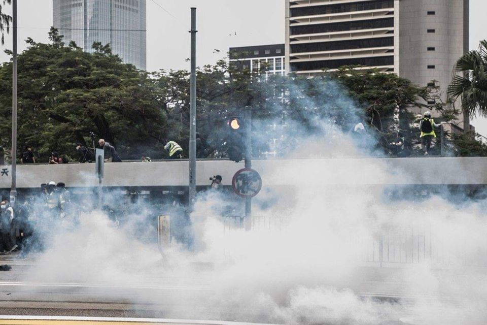 Tear gas rising from a street as protestors flee over a fence